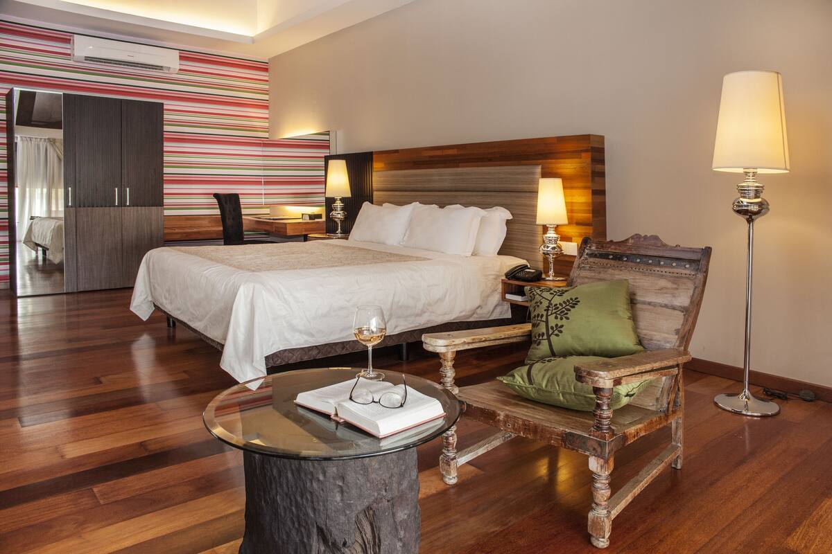 A hotel room with a large bed, a wooden chair, a round glass table, and a lamp.