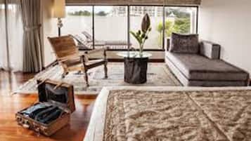Habitación amplia con una cama grande, un sofá, una silla de madera y una mesita con una planta. Se ve el exterior a través de una ventana.