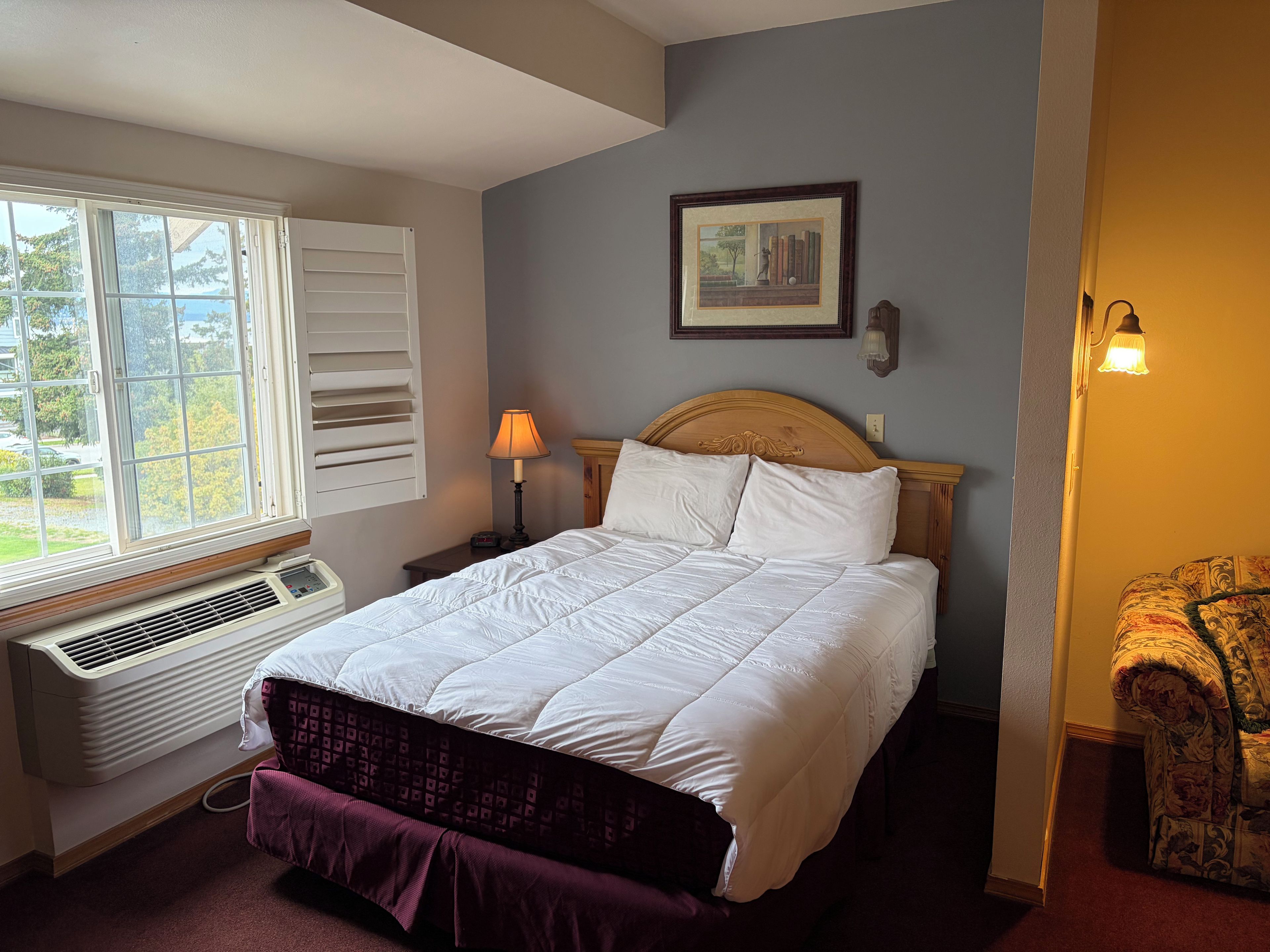 A bedroom with a bed, a window, a wall-mounted picture, a nightstand, a lamp, and a chair.