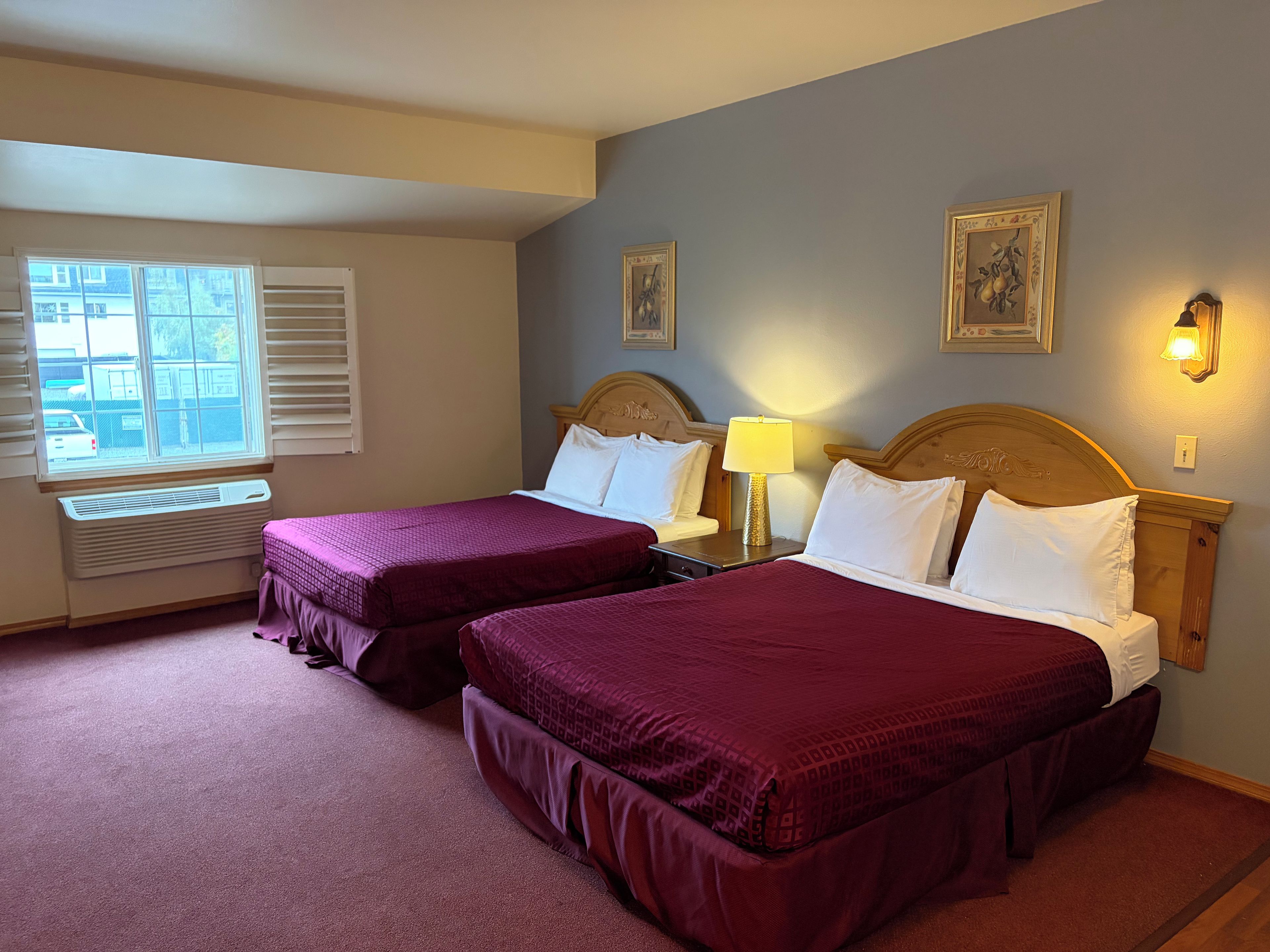A hotel room with two beds, a window with blinds, a wall-mounted lamp, and a framed picture.