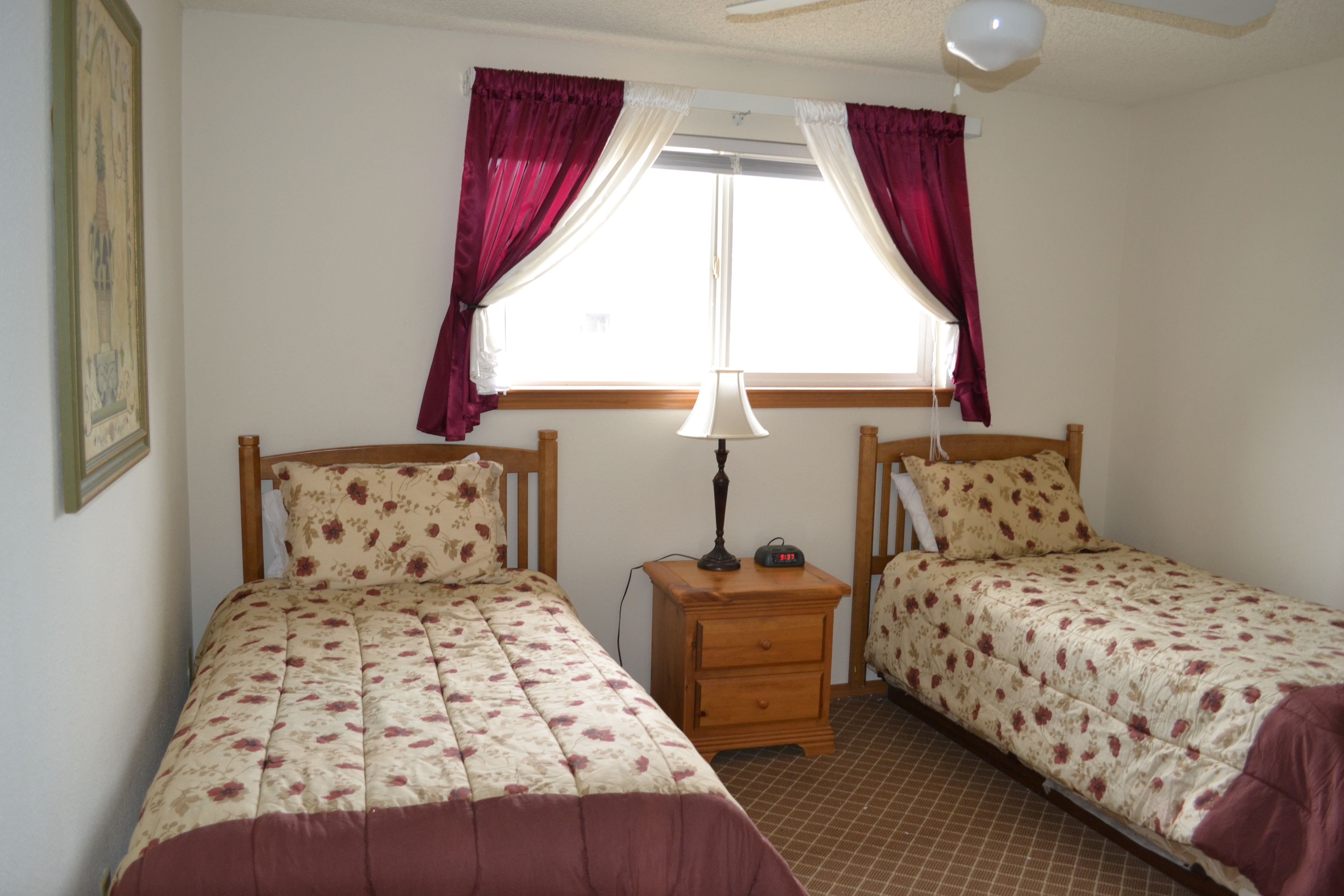 A bedroom with two single beds, a nightstand with a lamp, a window with curtains, and a ceiling fan.