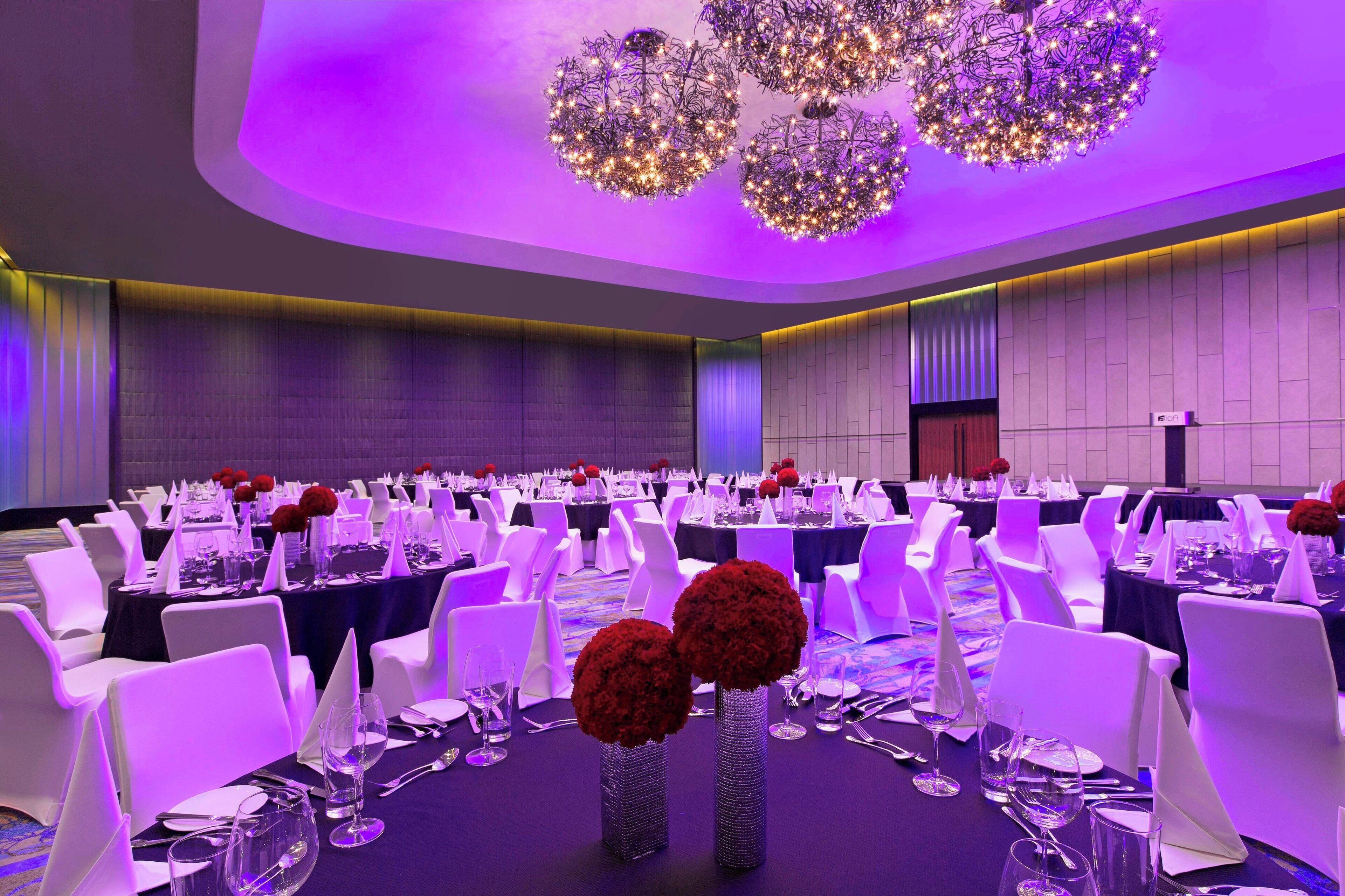 Un salón de banquetes con mesas redondas, sillas blancas e iluminación morada.