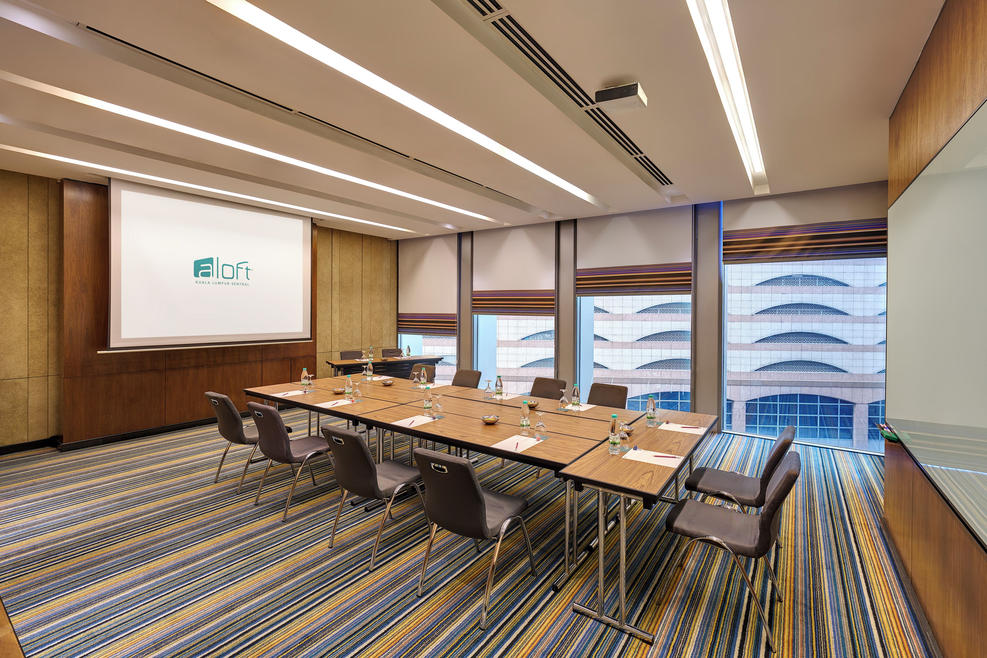 Une salle de conférence avec une longue table, des chaises, un écran de projection et une vue sur un bâtiment à travers de grandes fenêtres.