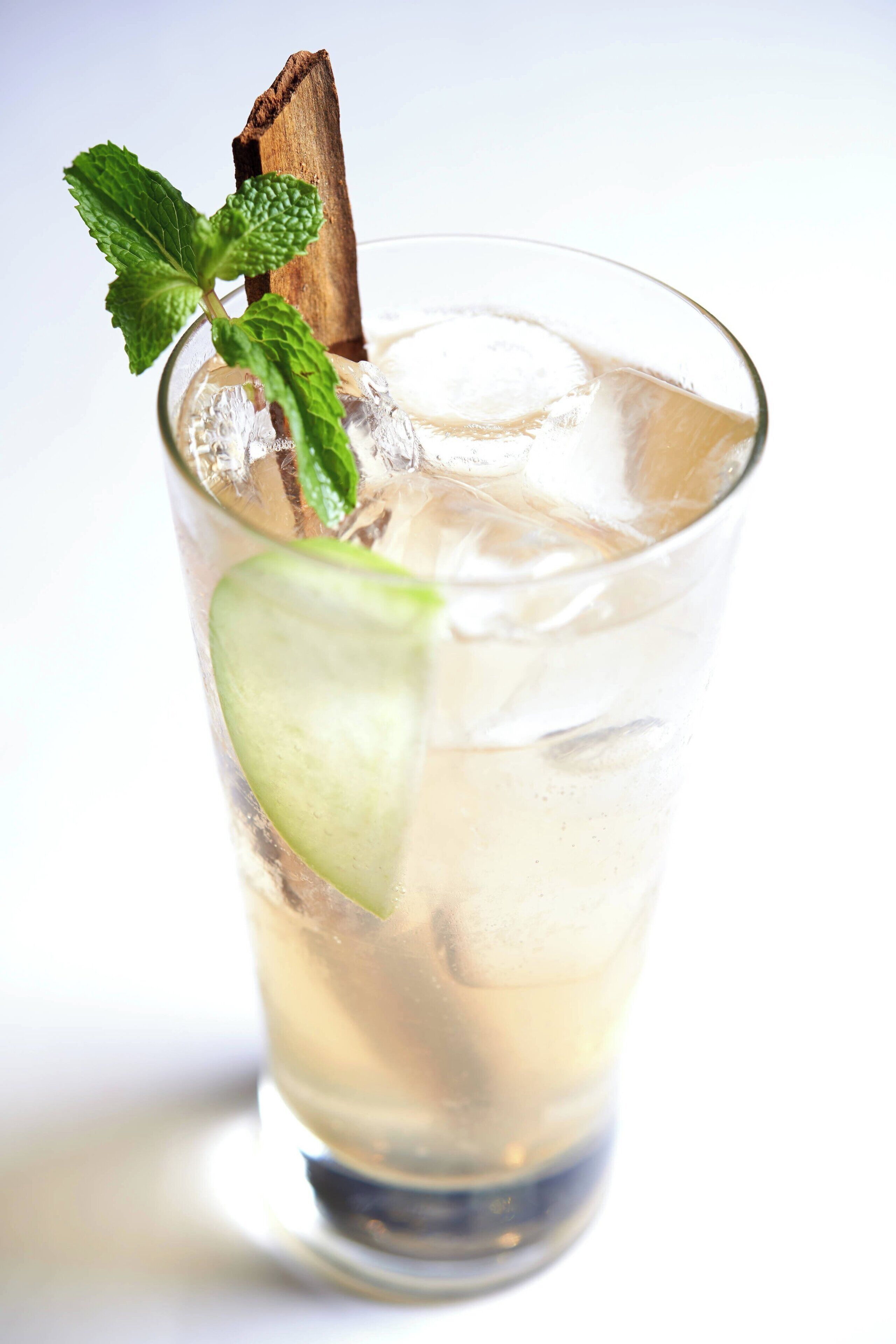 Un verre transparent contenant une feuille de menthe et une petite brochette en bois, et dans lequel on voit une boisson claire, de couleur pâle, avec des glaçons.