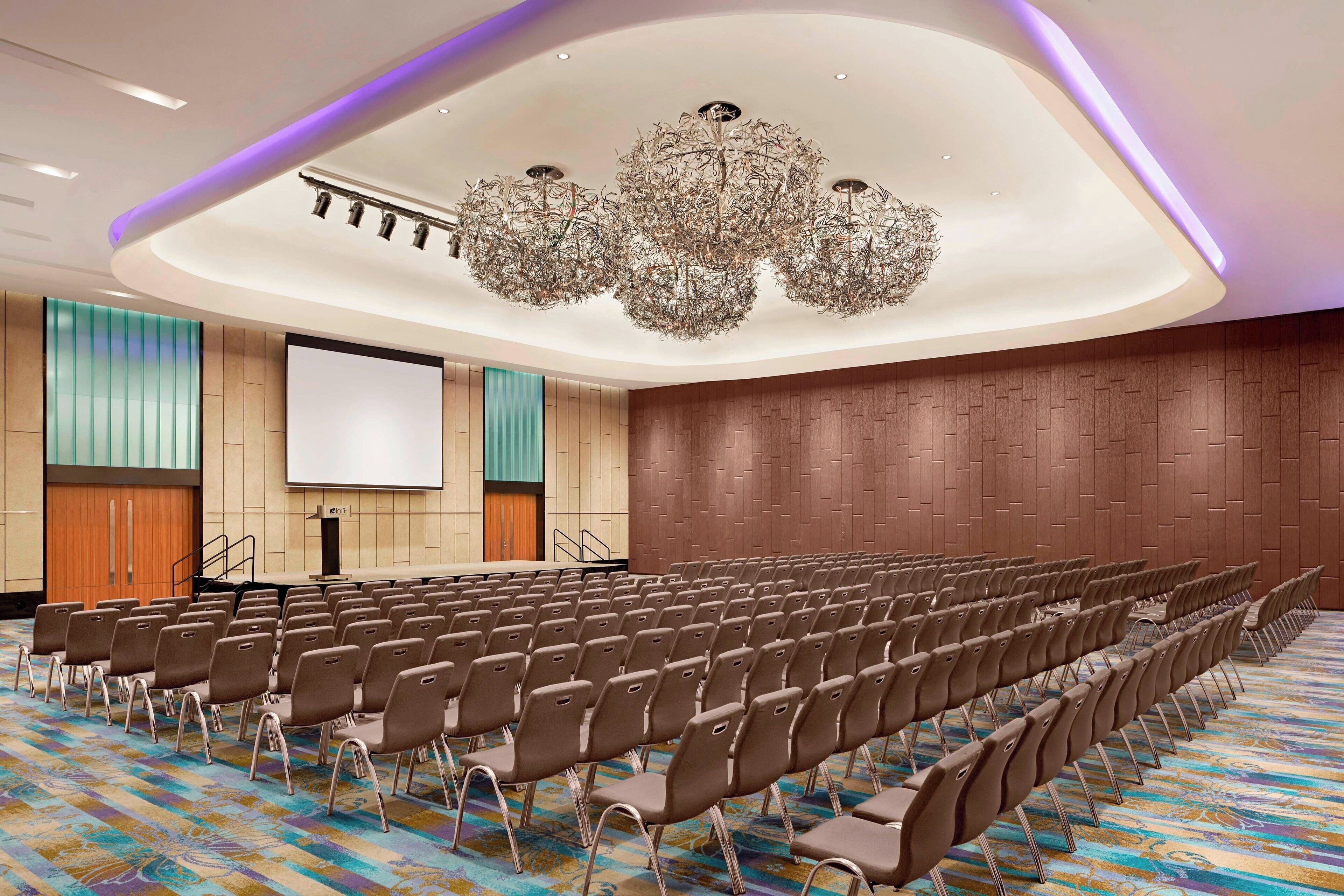 A conference room with rows of chairs, a large screen, and a modern chandelier.