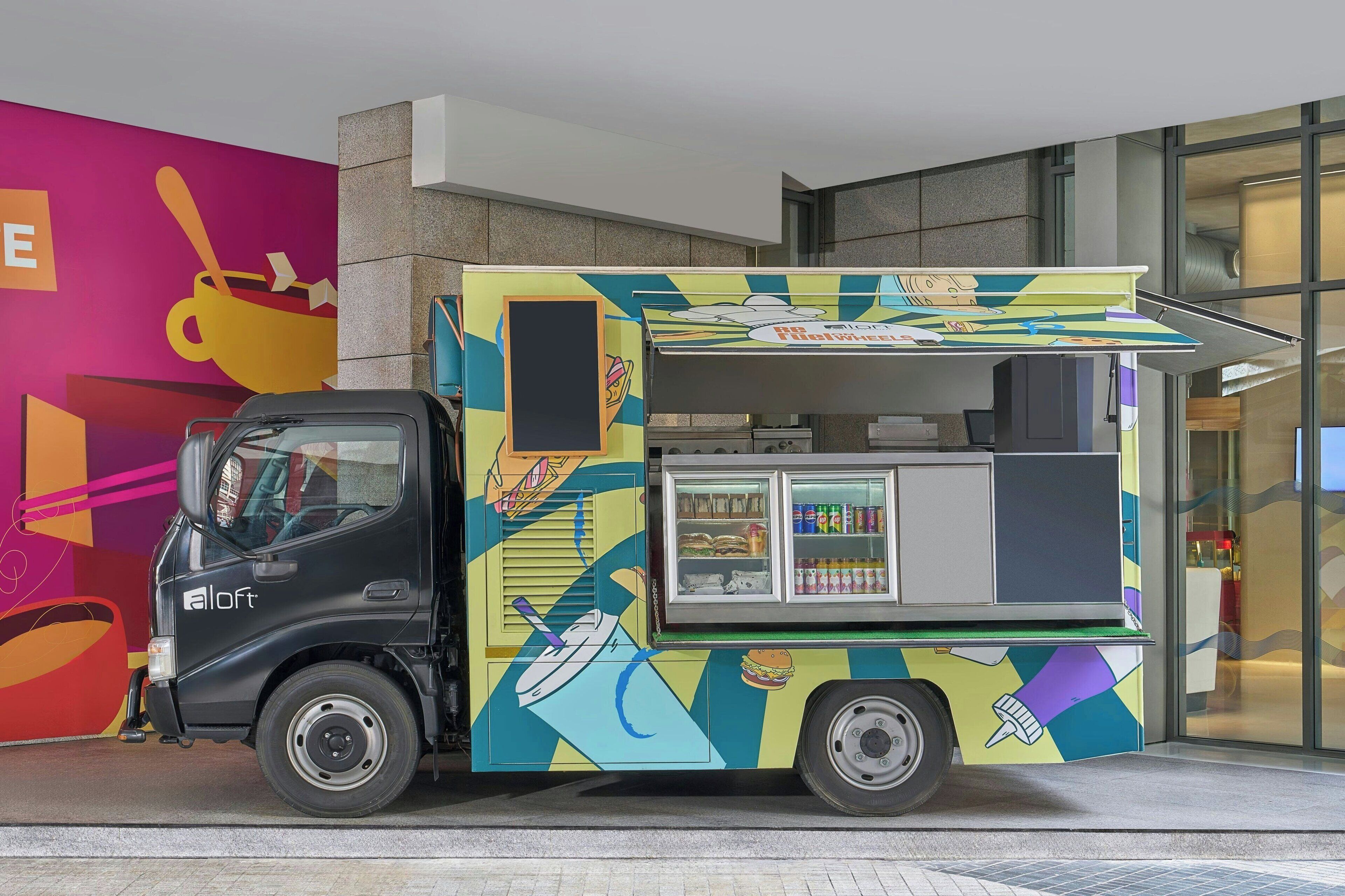 A colorful food truck with a black cab parked in front of a building with a vibrant mural.