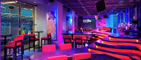 Zona de bar con asientos rojos y negros, una barra con un letrero de neón y un mural en la pared.