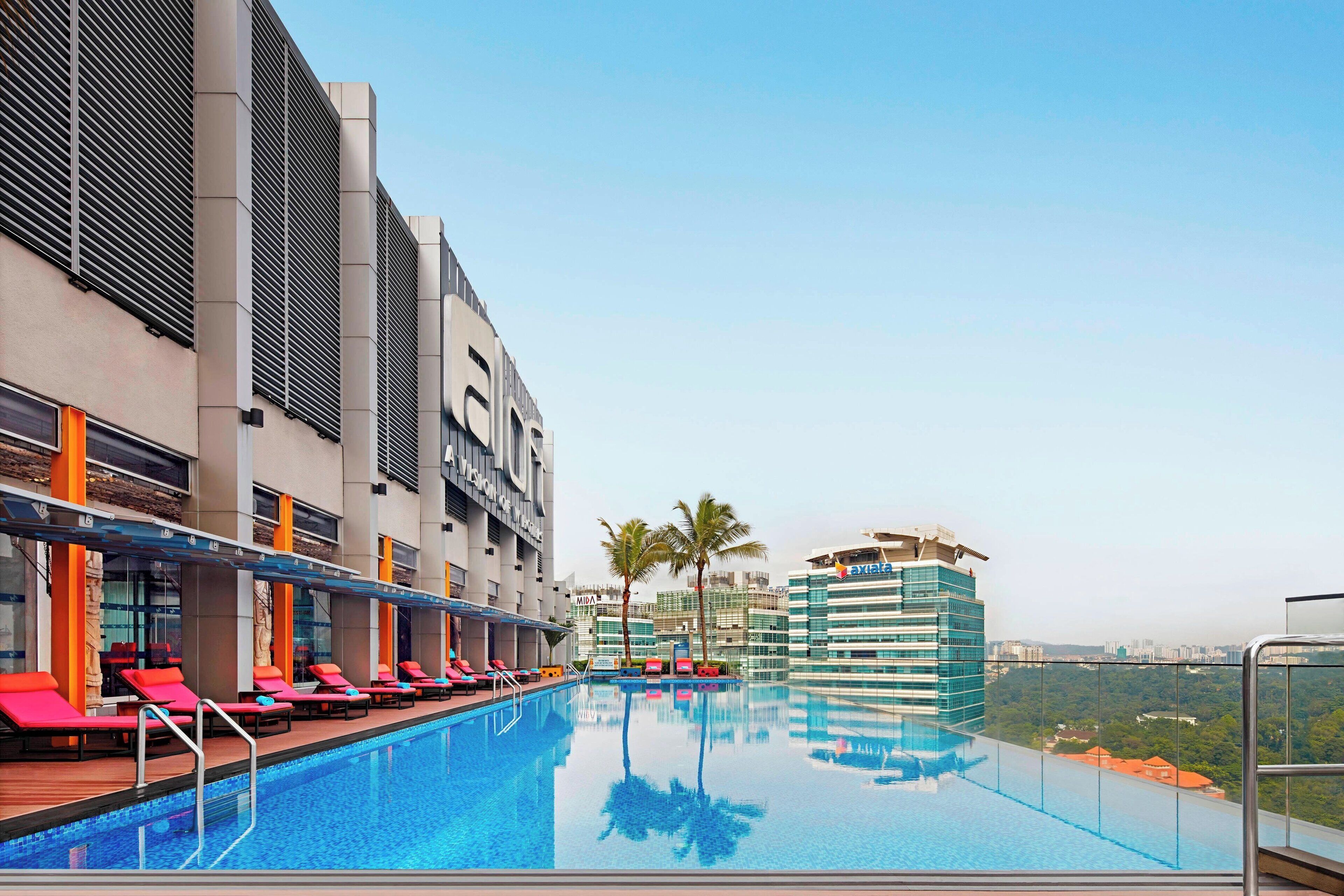 A rooftop infinity pool with red lounge chairs, a glass railing, and a view of a cityscape.
