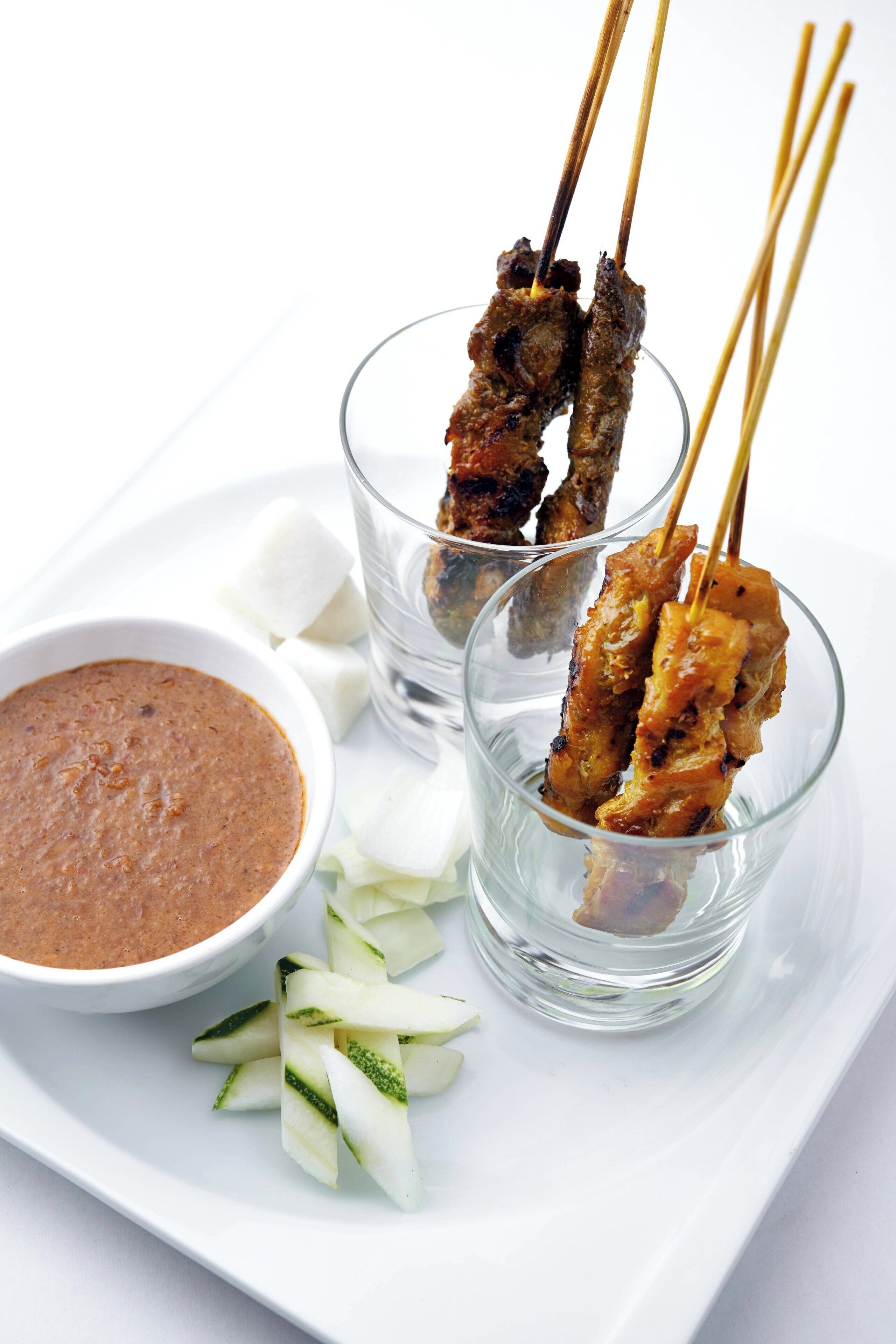 Skewers of grilled meat with a dipping sauce and cucumber slices on a white plate.