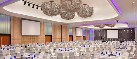 A large banquet hall with round tables and white chairs, a modern chandelier, and a large screen on the wall.