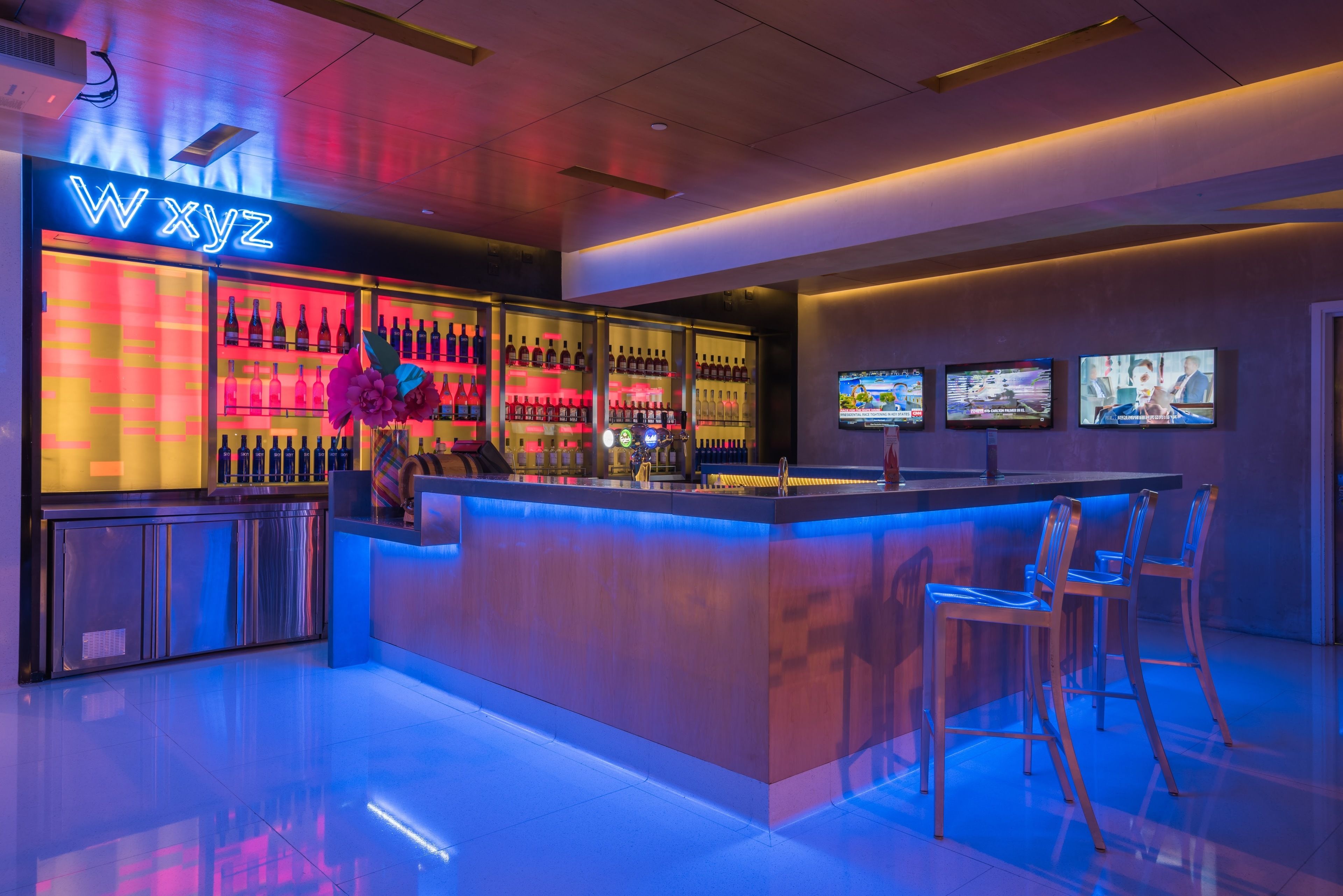 Uma área de bar moderna com um letreiro de neon, prateleiras cheias de garrafas e banquetas.