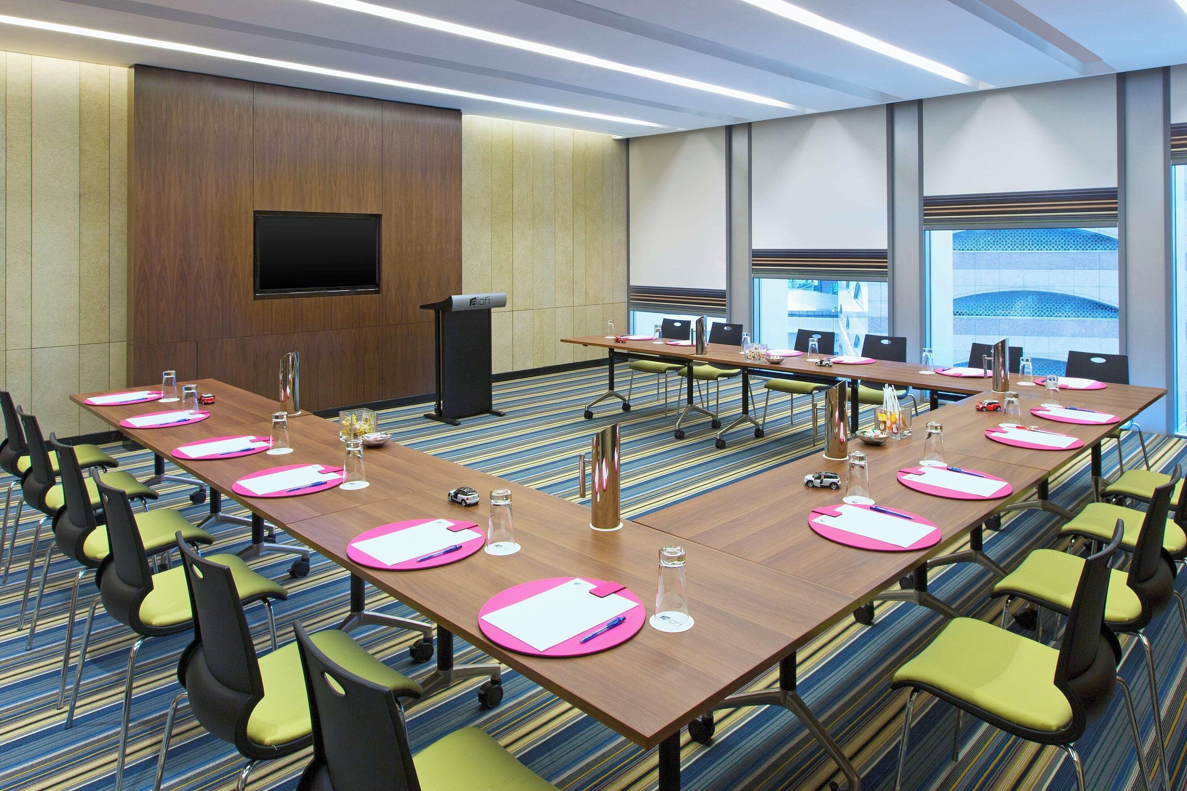 Un salón de conferencias con una larga mesa, sillas, un atril y una pantalla de televisión.