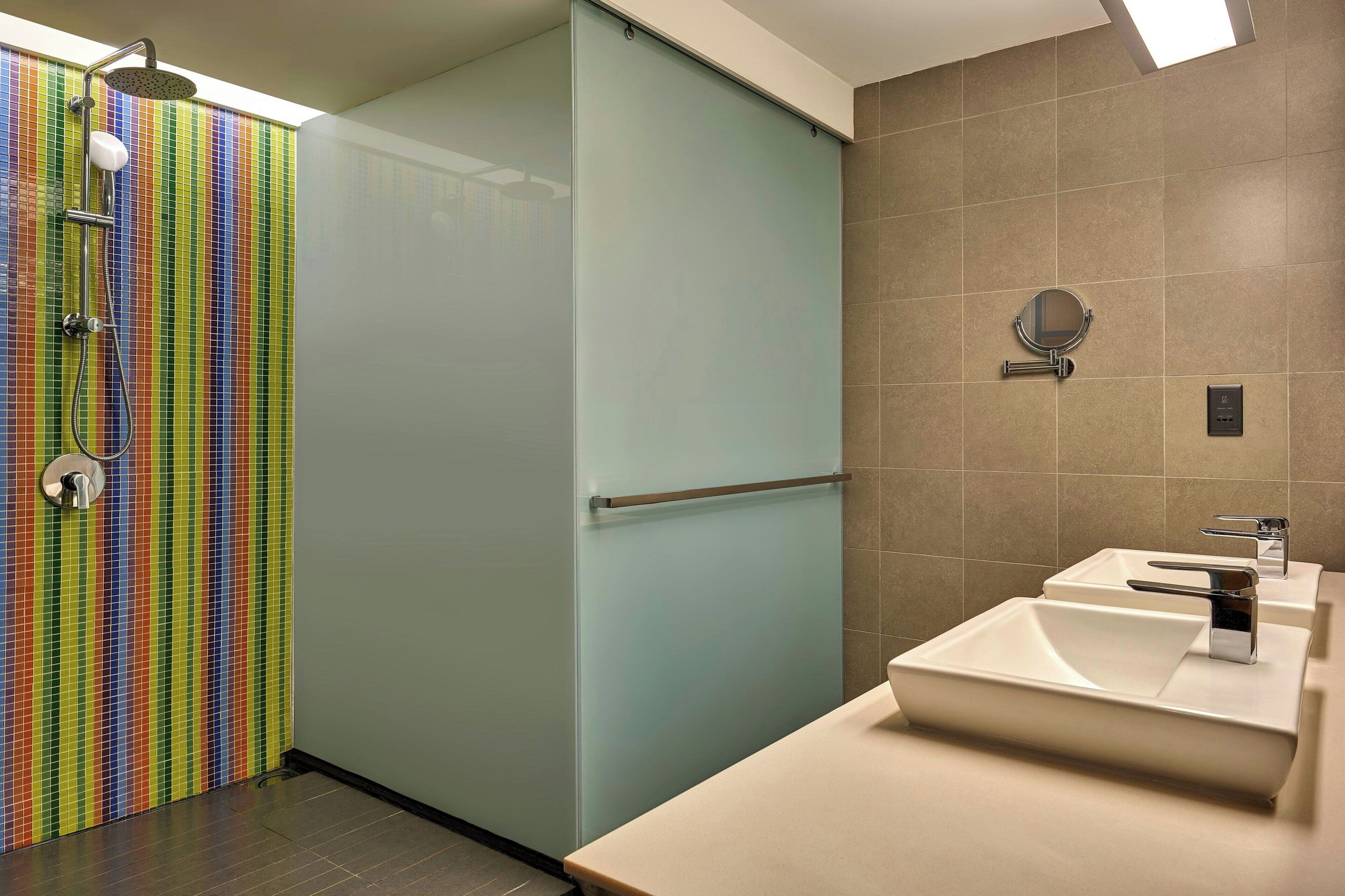 Ein Badezimmer mit farbenfroher gefliester Duschwand, einer Milchglas-Tür, einem beigen Waschbecken mit modernem Wasserhahn und einem kleinen Spiegel an der Wand.