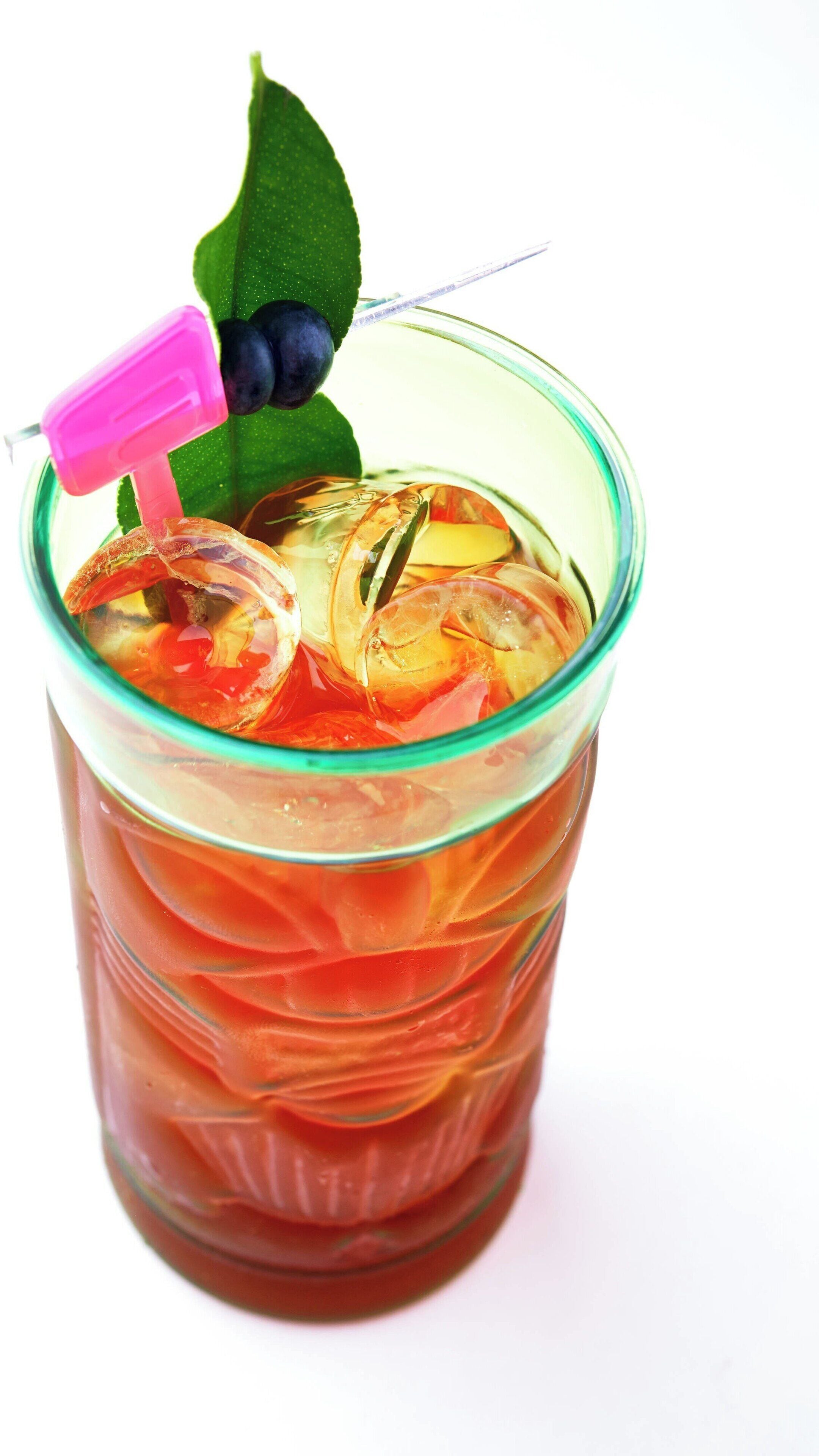 A glass with a pink straw, containing a red beverage and ice cubes, with a blueberry and a leaf garnish.