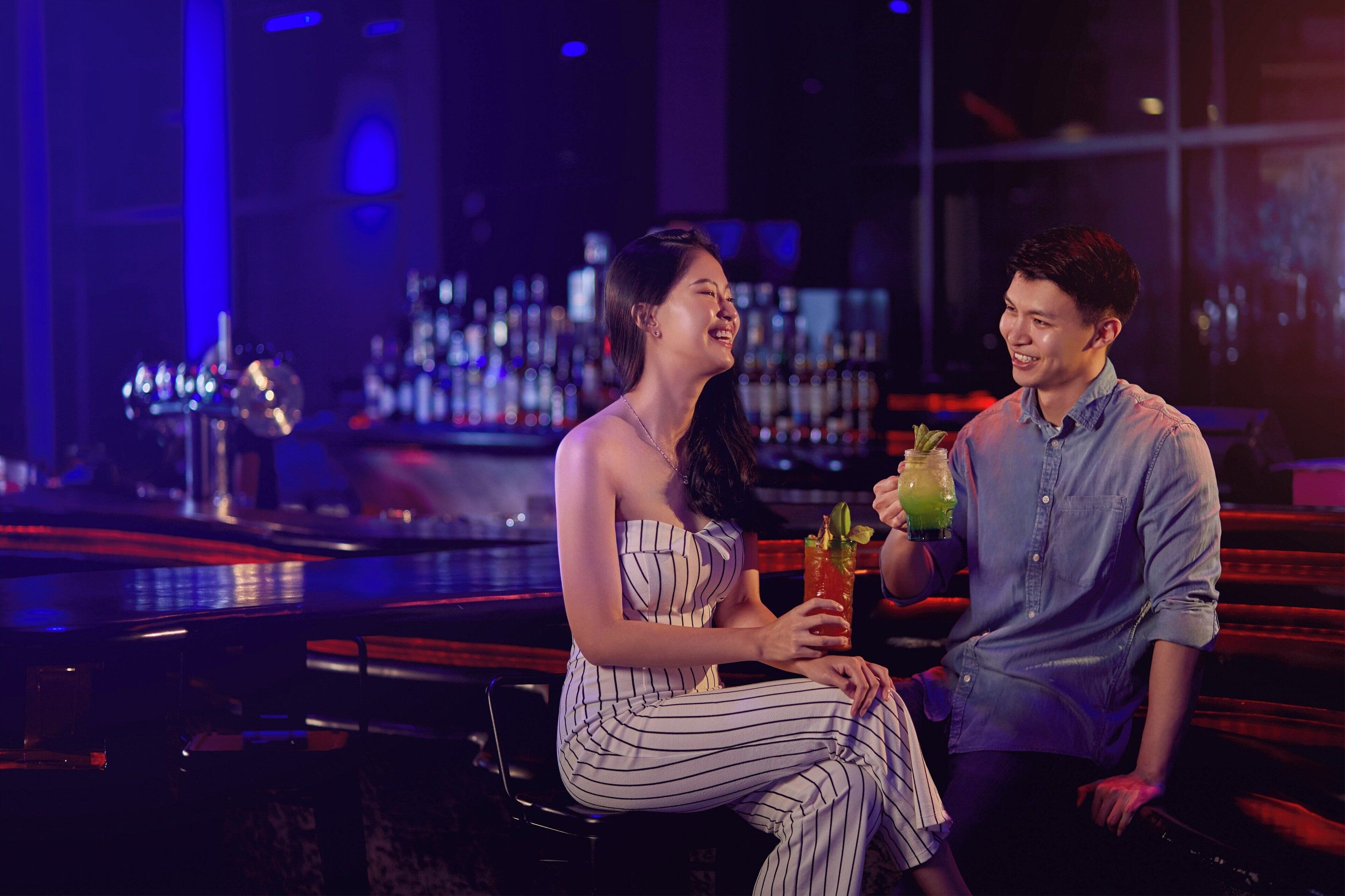 A bar setting with a person seated and another standing, holding a drink.