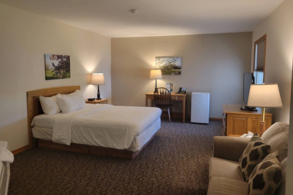 A hotel room with a bed, two bedside tables with lamps, a desk with a chair, a small refrigerator, a sofa, and two wall-mounted lamps.