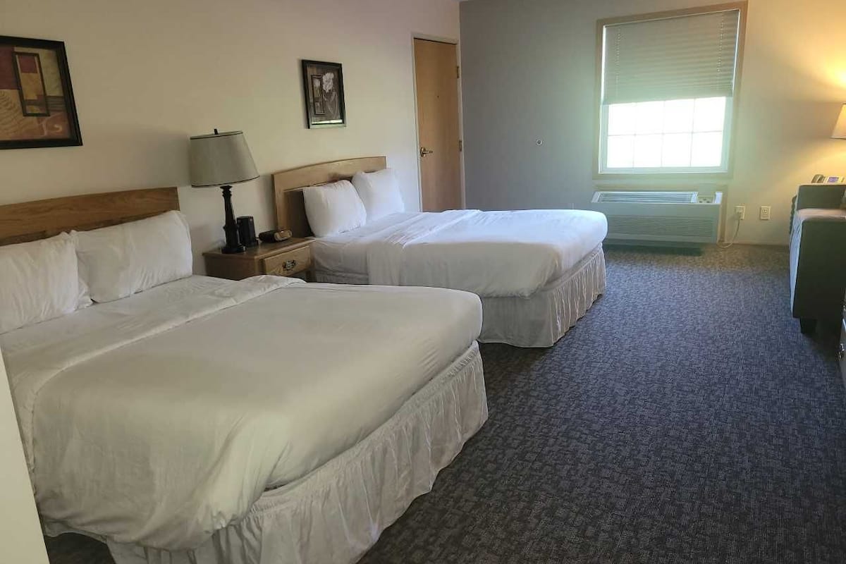 A hotel room with two single beds, a nightstand with a lamp, a dresser, and a window with blinds.