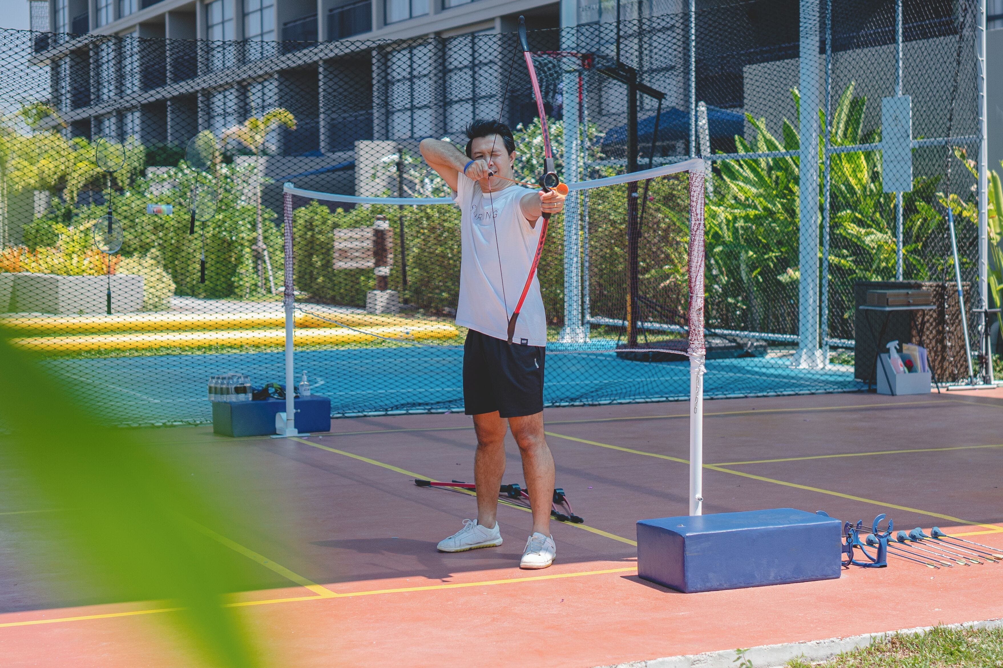 A person practicing archery on an outdoor court with a netted enclosure, benches, and a building in the background.
