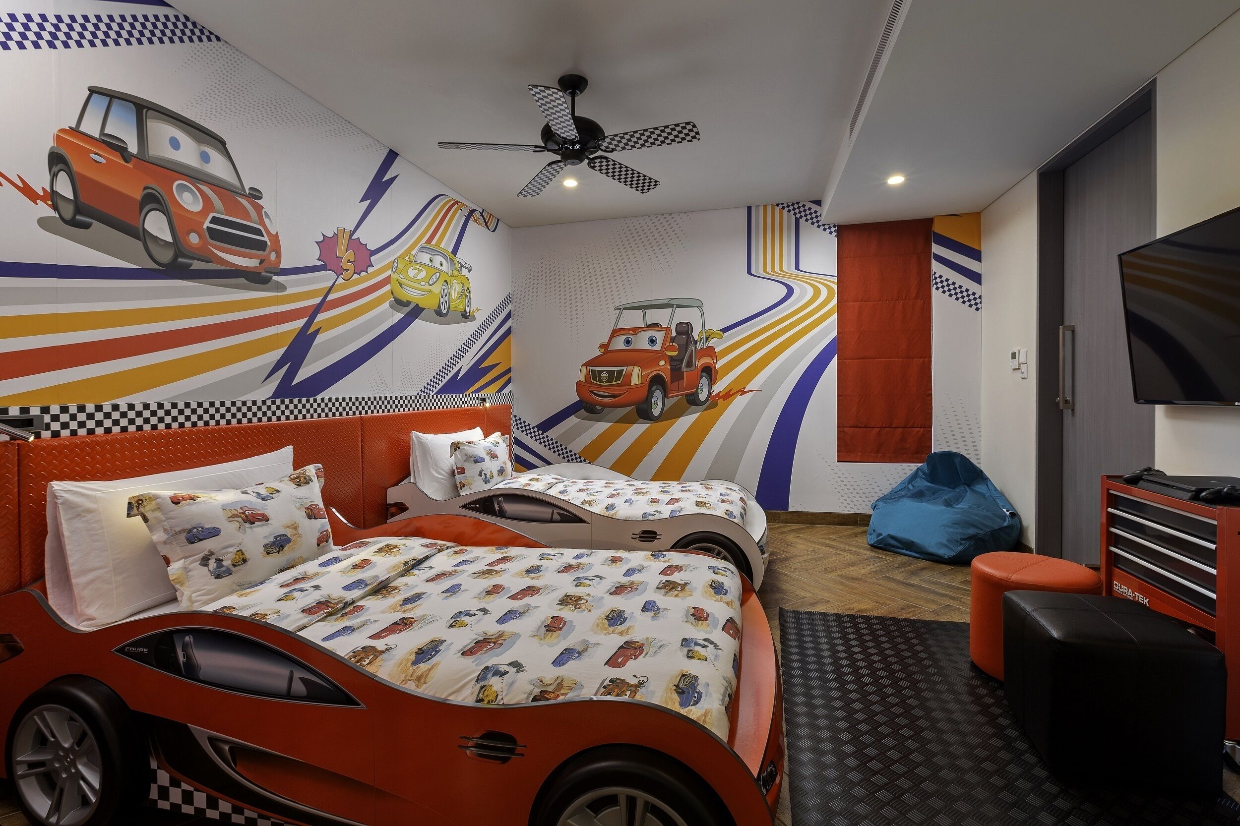 Habitación infantil con temática de carreras, que incluye una cama, dos camas individuales, un ventilador de techo, un televisor y un sillón de peluche.