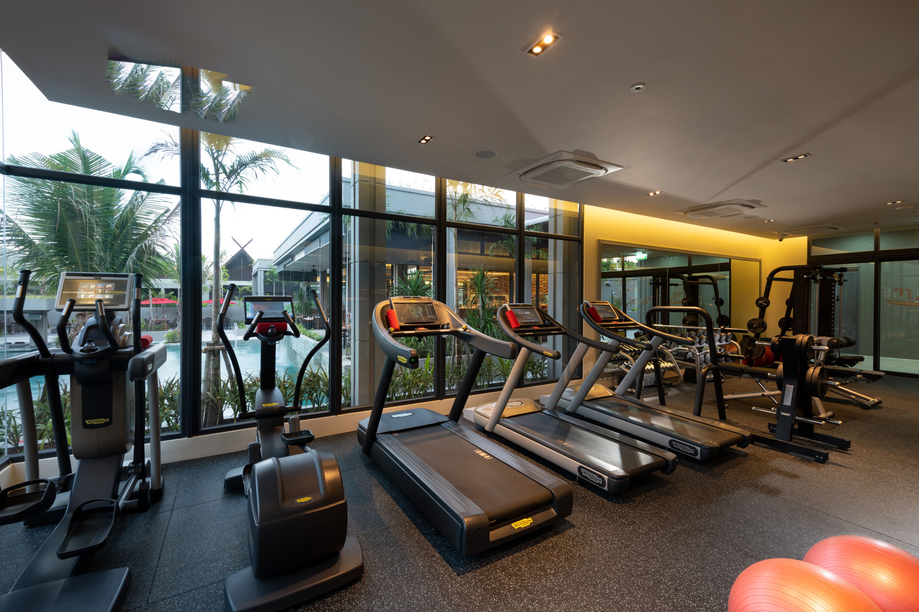 Una palestra con tapis roulant, ellittiche e palle da ginnastica, ampie finestre vista palme e piscina.