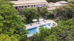 Un edificio grande con varias habitaciones, rodeado de abundante vegetación y una piscina con sillones de descanso.
