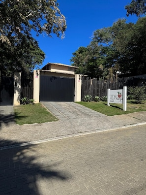 A gated property entrance with a paved driveway, a signboard, and a fenced area with greenery. - Vila Pitanga (Búzios)