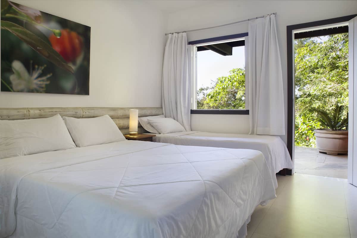 Habitación de hotel con dos camas, un ventanal con cortinas, una planta en maceta y un cuadro en la pared.