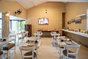 A dining area with white tables and chairs, a buffet with various food items, and a television screen on the wall. - Vila Pitanga (Búzios)