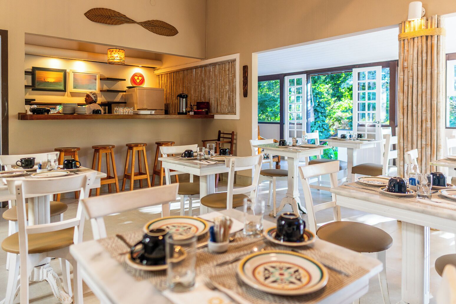 A restaurant with white furniture, a kitchen area, and large windows with a view of greenery.
