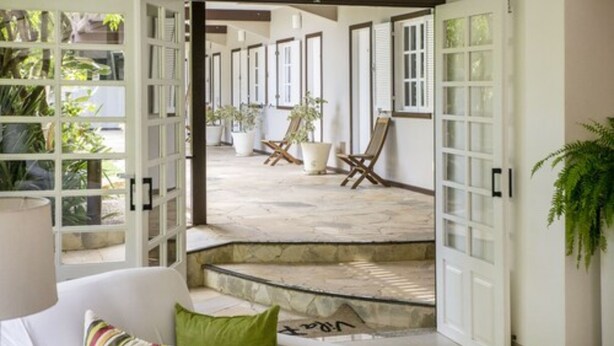 Amplia zona de patio con un sofá blanco, una almohada verde y una silla de madera. Hay plantas en macetas y una puerta blanca que da a otra habitación.