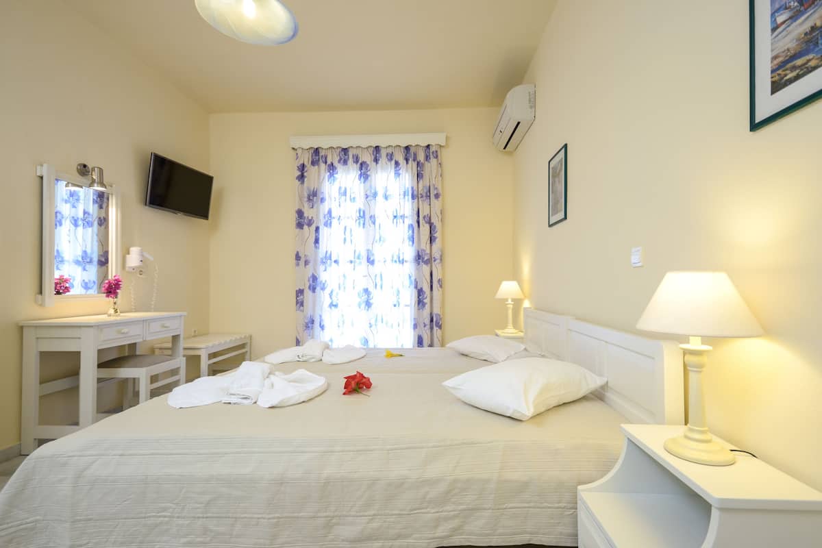 A hotel room with a bed, bedside tables, a TV, a window with curtains, and an air conditioning unit.