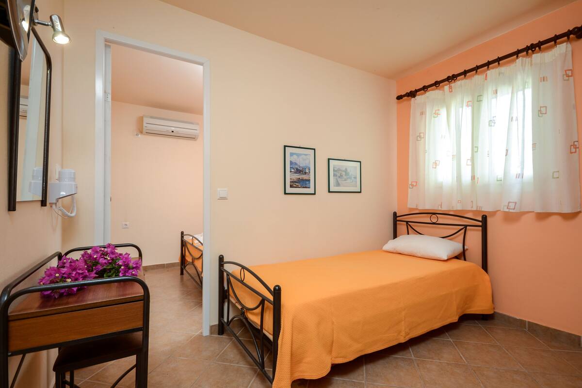 A bedroom with a single bed, a bedside table with a flower arrangement, a chair, and a window with curtains.