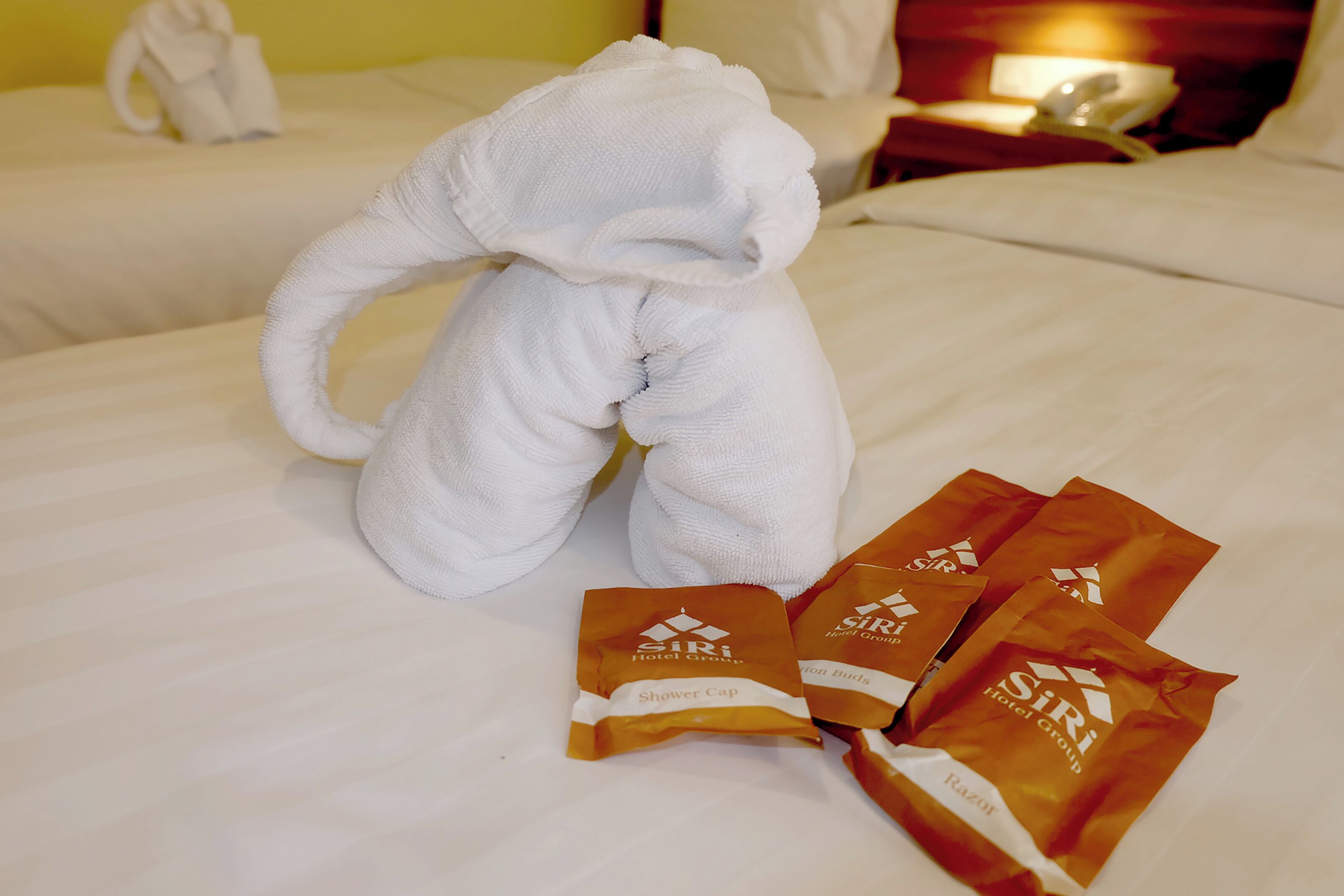 Ein Bett mit einem weißen Elefanten-Handtuch, einer Reihe brauner Taschen mit der Aufschrift "Siri Hotel Group“ und einem weißen Handtuch auf dem Bett.