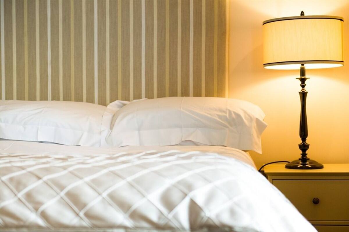 Un lit bien fait, avec du linge de lit blanc et une couette matelassée, une lampe de chevet sur une table de nuit, et un mur à rayures en arrière-plan.