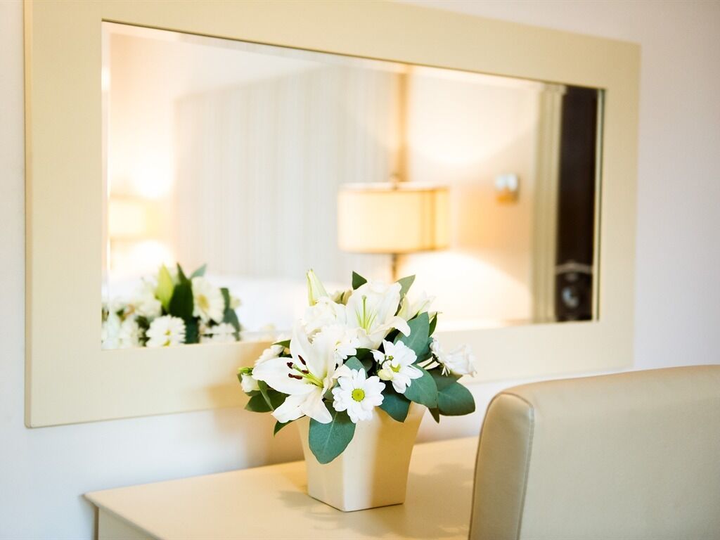 A mirror reflecting a room with a bedside table, a lamp, and a vase of white flowers.