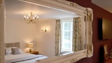 Quarto com um espelho grande e ornamentado, um lustre, uma cama com colcha estampada de flores e uma janela com cortinas florais.