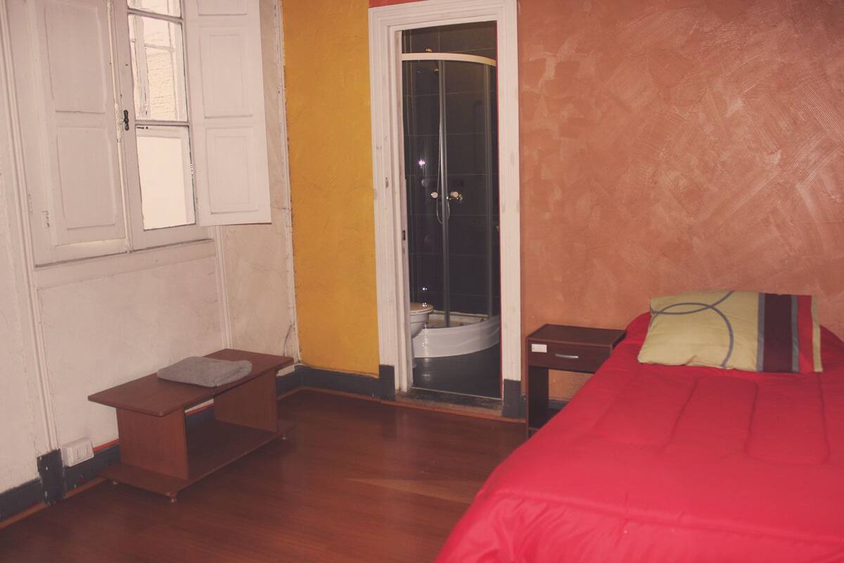 A bedroom with a wooden floor, a bed with a red blanket, a small wooden nightstand, a window with white shutters, and a bathroom with a shower and toilet visible through a glass door.