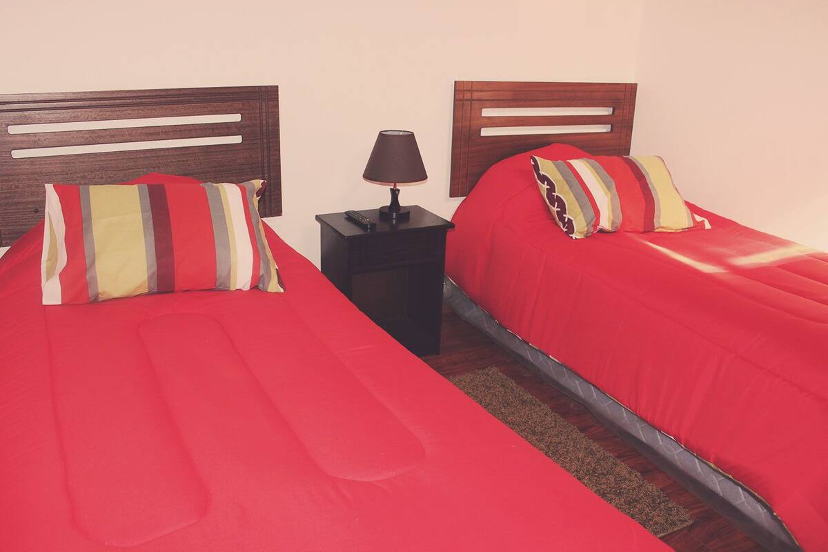 Two single beds with red bedding and striped pillows, a nightstand with a lamp, and a wooden headboard.