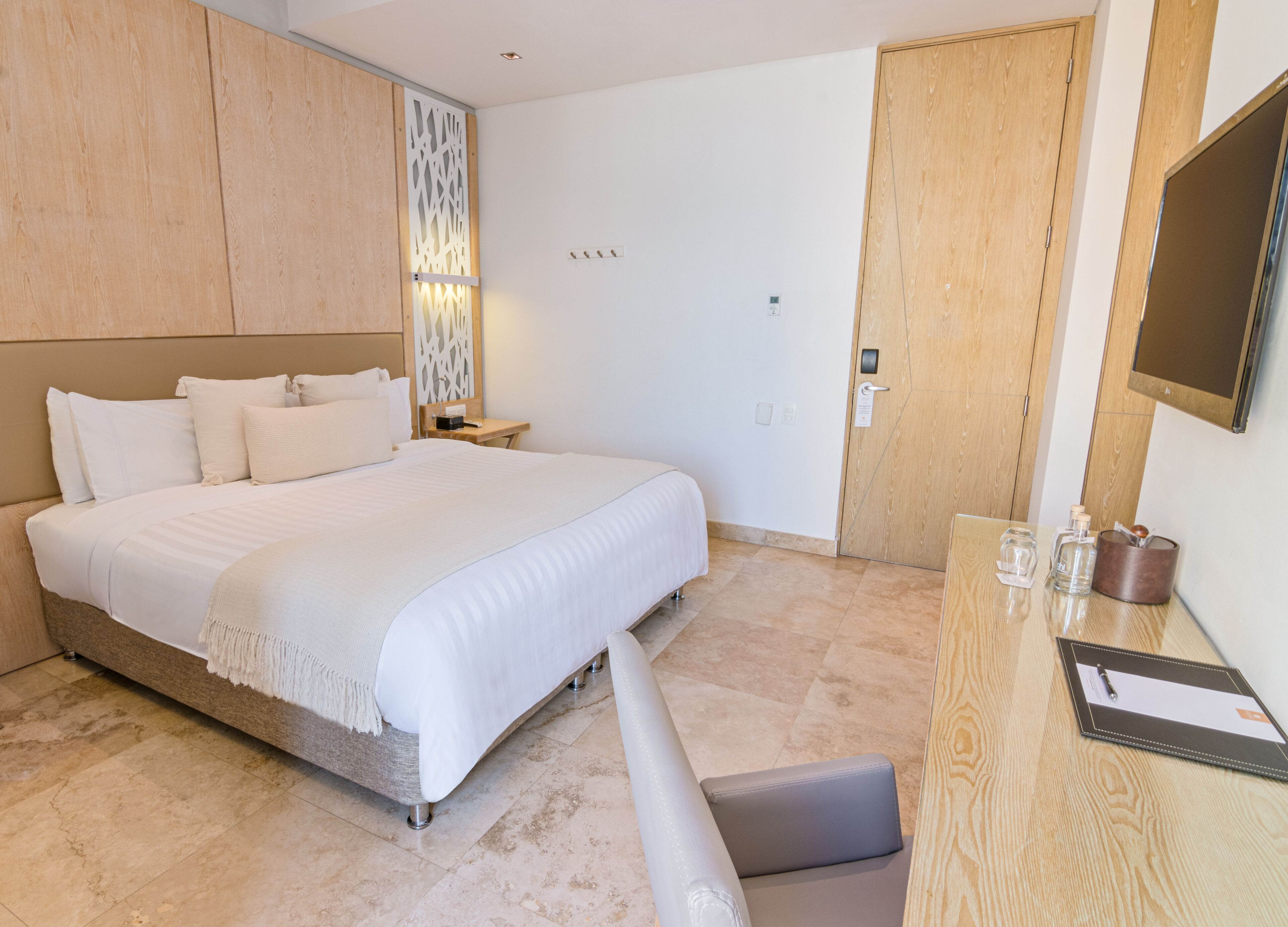 A modern hotel room with a large bed, wooden headboard, a bedside table with a lamp, a desk with a chair, and a TV mounted on the wall.
