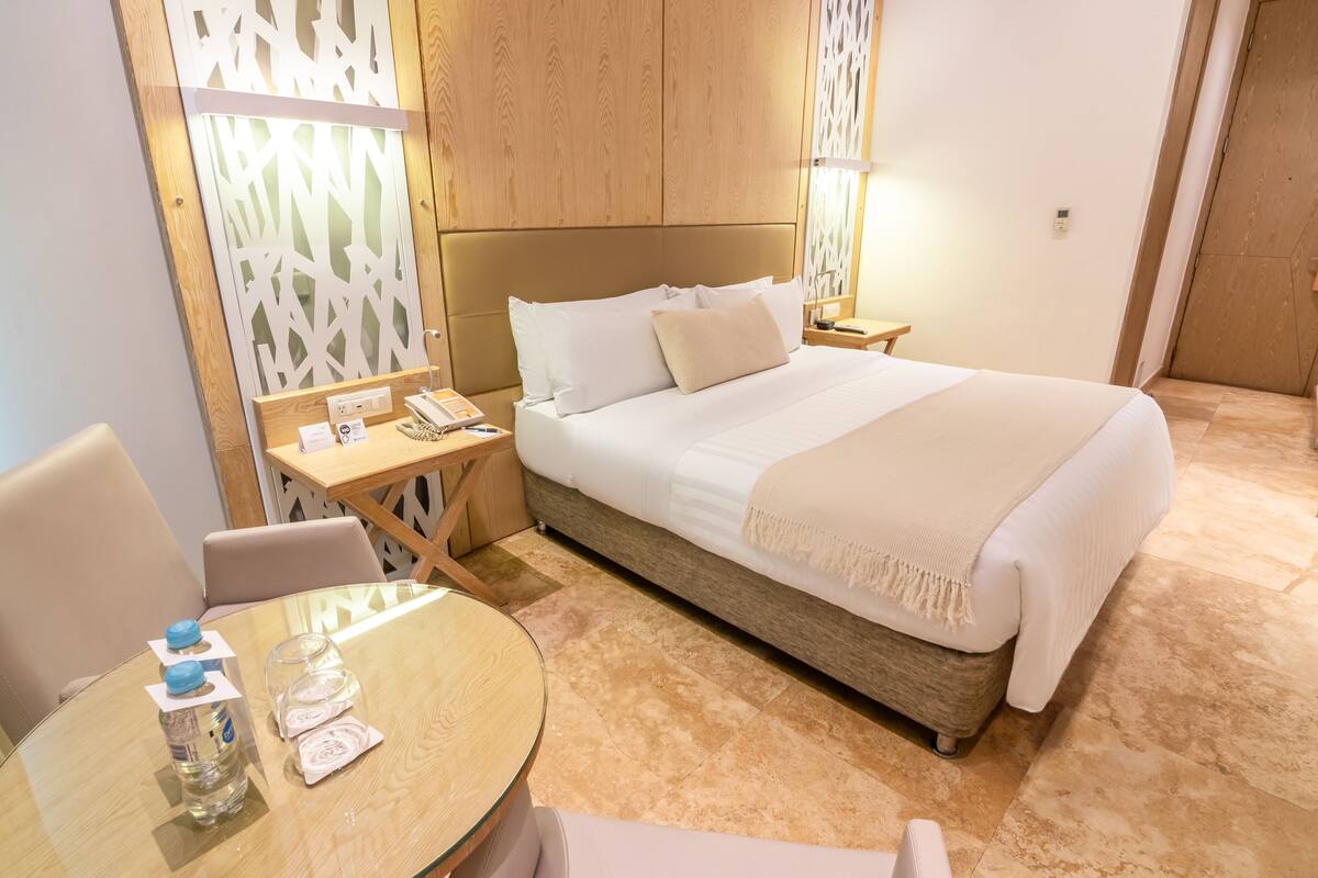 Habitación de hotel con cama, mesitas de noche, una mesa redonda con sillas y un panel decorativo en la pared.