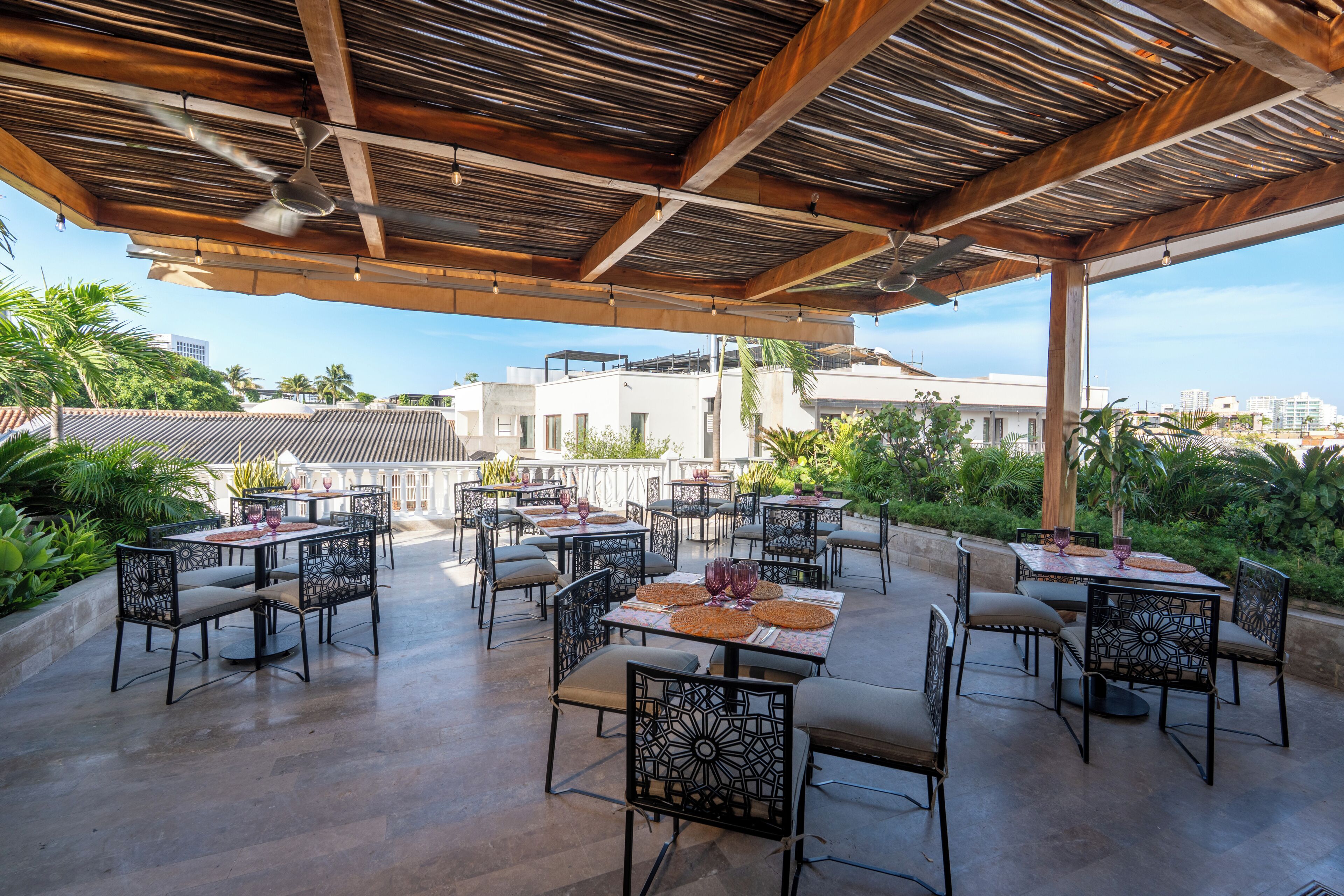 An outdoor patio with tables and chairs, a wooden ceiling, and a view of buildings in the background.
