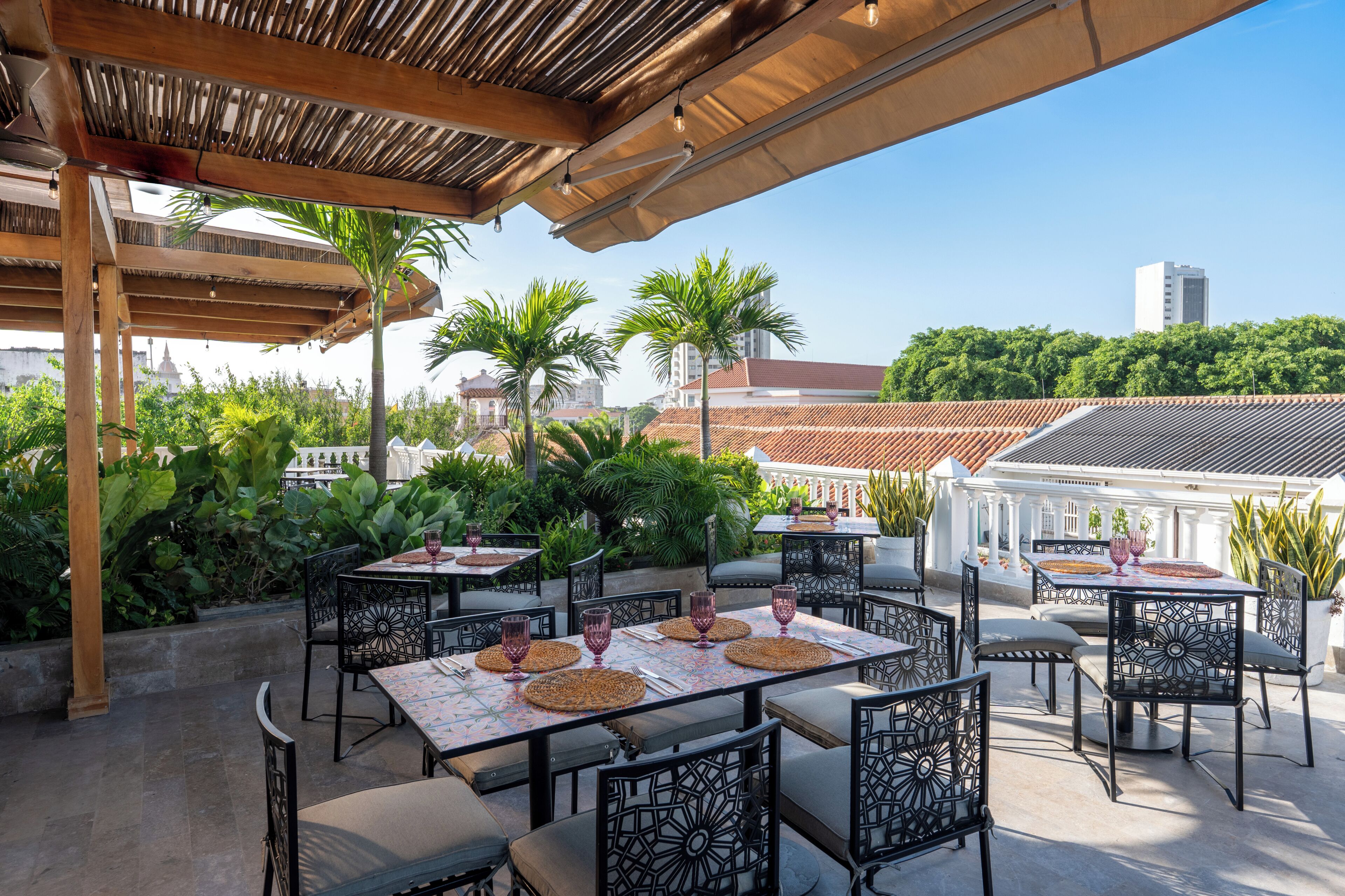 Un patio al aire libre con mesas y sillas, rodeado de vegetación y vistas a edificios.