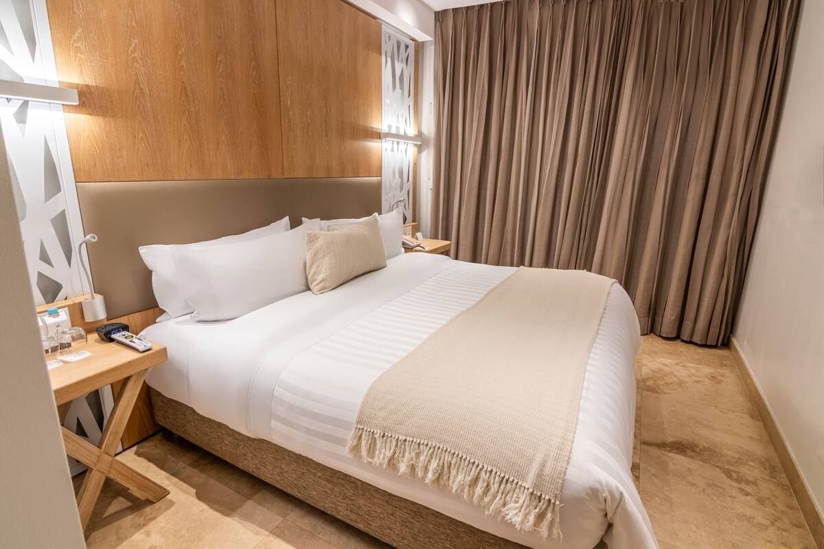 Una habitación de hotel con una cama grande, cabecero de madera, una mesita de noche con lámpara y una cortina.