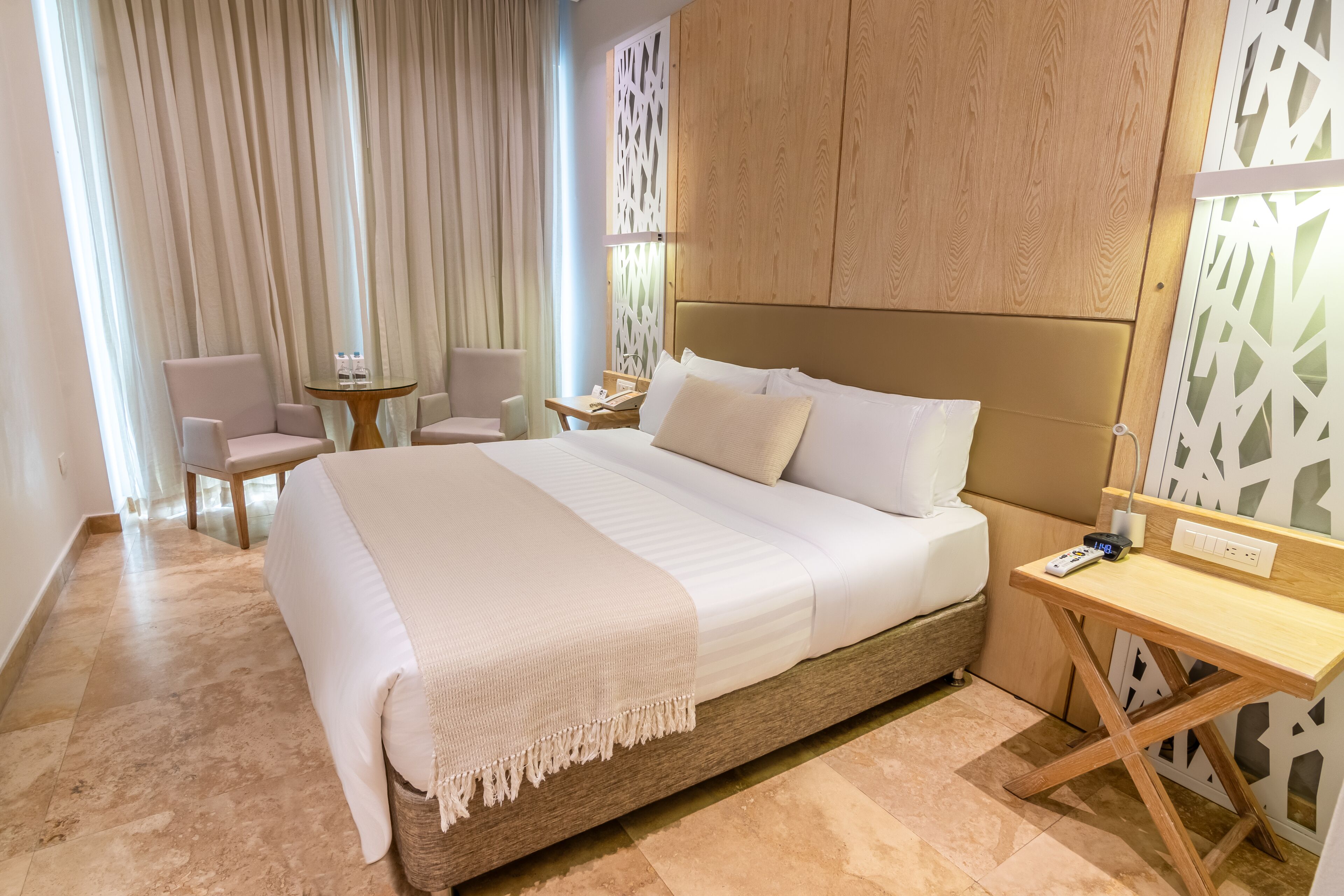 A modern hotel room with a large bed, a wooden headboard, a small desk, and a seating area with two chairs.