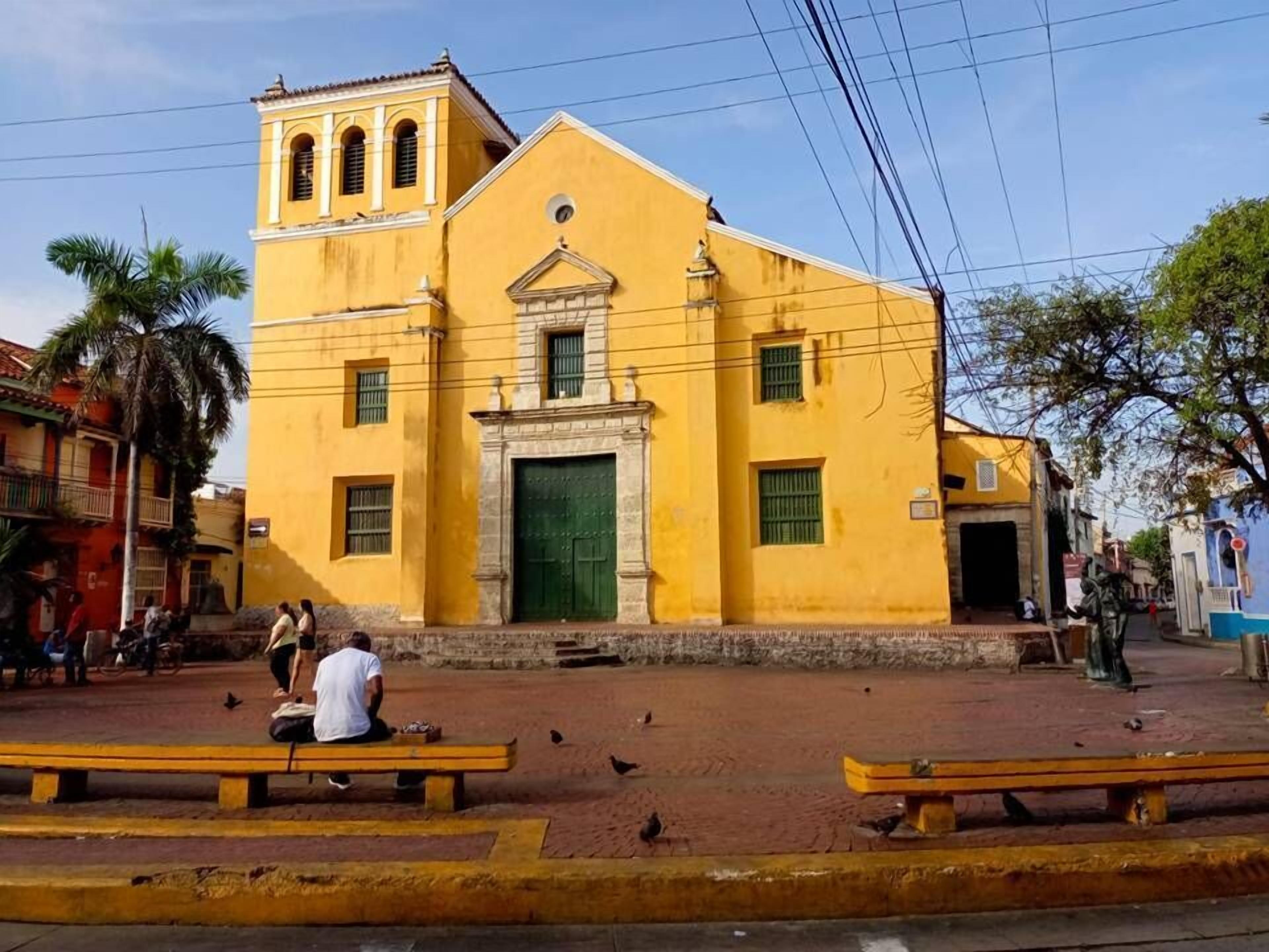 A yellow building with a green door and windows, surrounded by benches and people.