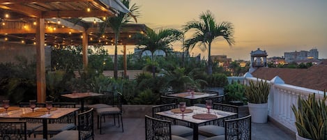 An outdoor dining area with tables and chairs, palm trees, and a view of a cityscape at dusk.