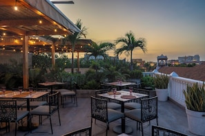 An outdoor dining area with tables and chairs, palm trees, and a view of a cityscape at dusk.