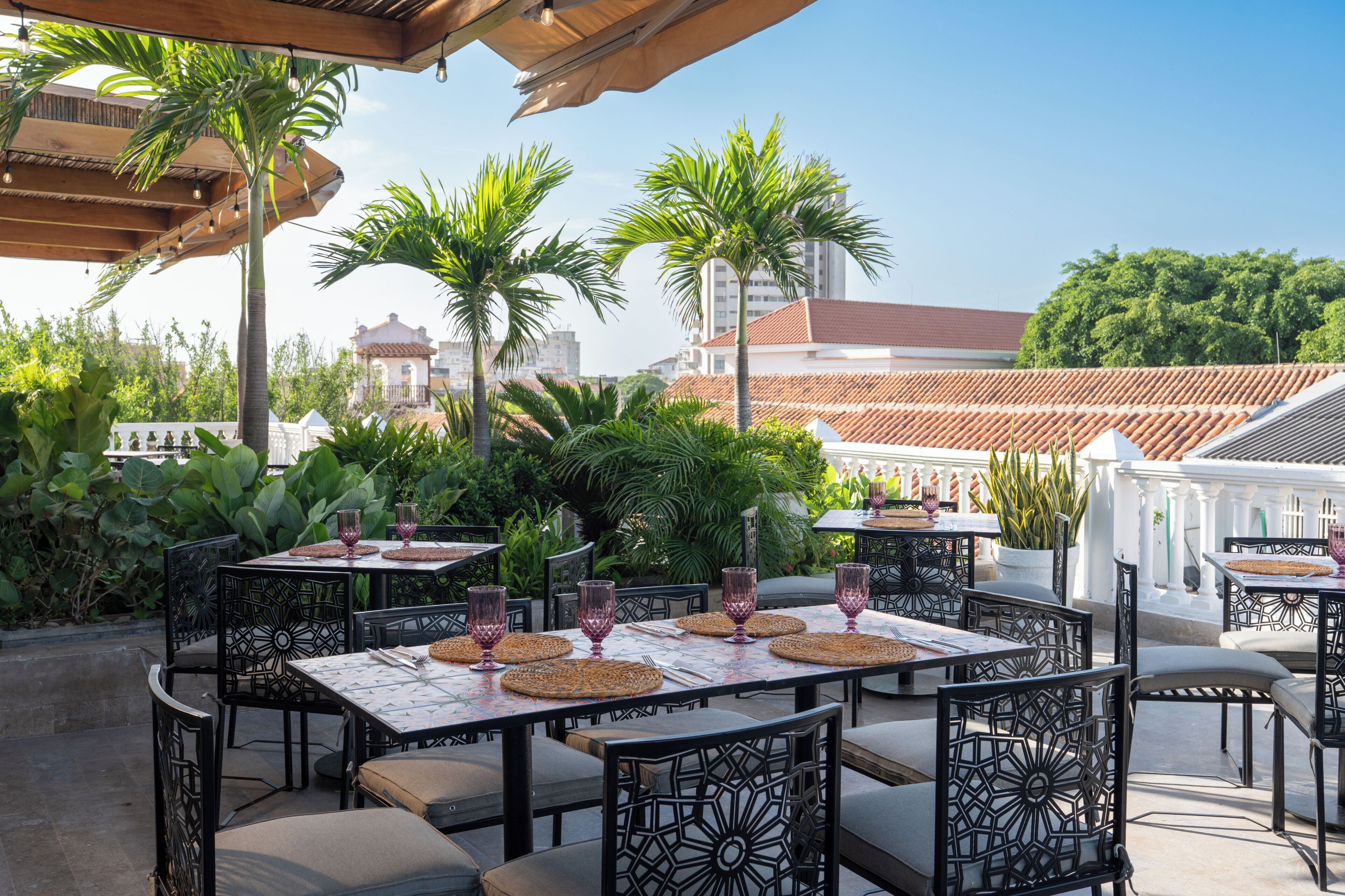 An outdoor dining area with tables and chairs, surrounded by greenery and buildings in the background.
