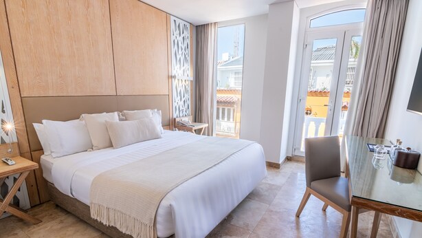 Une chambre d’hôtel avec un grand lit, un bureau, une chaise et un balcon donnant sur des immeubles.