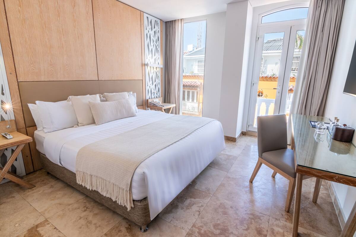 Habitación de hotel con una cama grande, un escritorio, una silla y un balcón con vistas a edificios.